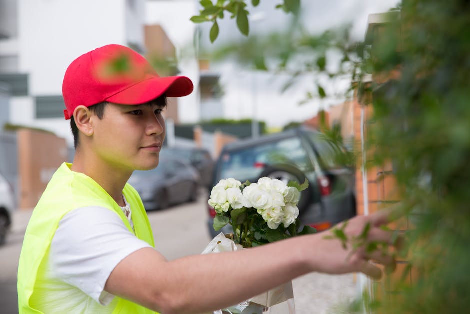 A young male flower delivery courier, wearing a red cap and a high-visibility yellow vest over a white t-shirt, is seen outdoors on a street in Kentish Town. He is holding a bouquet of fresh white hyd