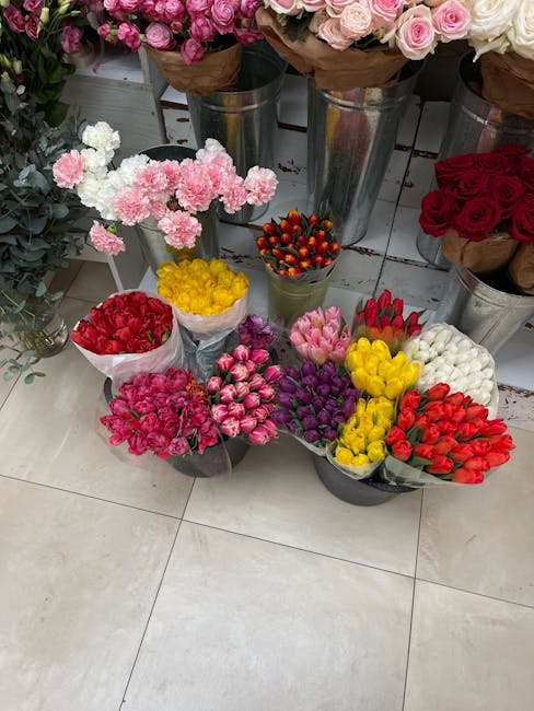 A display of various fresh flower bouquets and floral arrangements arranged on a tiled floor in front of metal vases filled with larger flower bouquets. The smaller bouquets feature tightly clustered,