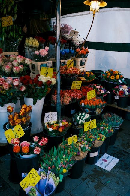 A vibrant outdoor flower stall displaying a variety of fresh floral arrangements and bouquets in black, white, and clear plastic buckets. The flowers include pink and white roses, orange tulips, yello
