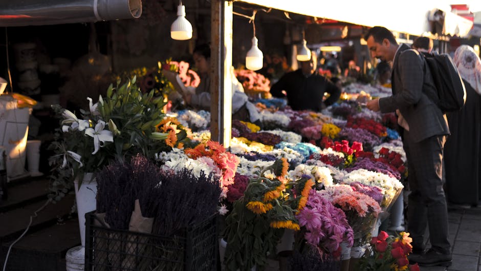 A vibrant outdoor flower stall with a variety of floral arrangements and potted plants. In the foreground, there are lush red and green flowering plants, possibly begonias, with numerous small red blo
