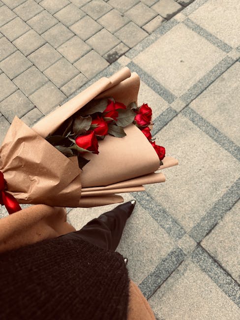 A hand wearing a floral-patterned glove holds a bouquet of fresh flowers wrapped in light pink paper, with pink blossoms visible, ready for flower delivery. The bouquet is being positioned on a dark b