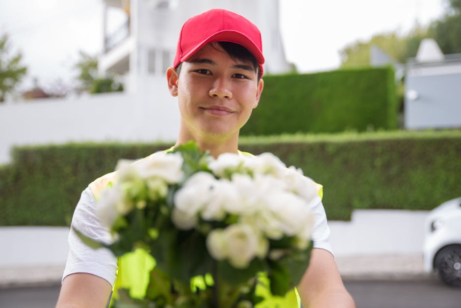A delivery person wearing a bright yellow high-visibility vest and a red cap holds out a bouquet of fresh white flowers towards the camera. The bouquet features a variety of white blooms with green fo