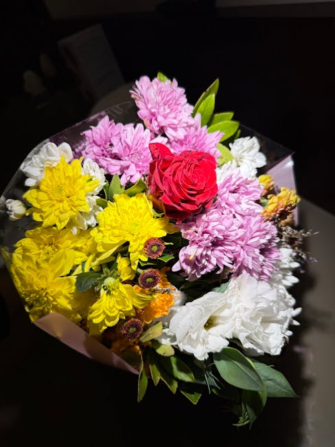 A vibrant bouquet of fresh flowers arranged with a variety of blooms including a large pink chrysanthemum, bright yellow chrysanthemums, a prominent red rose, white lilies, and smaller purple and oran