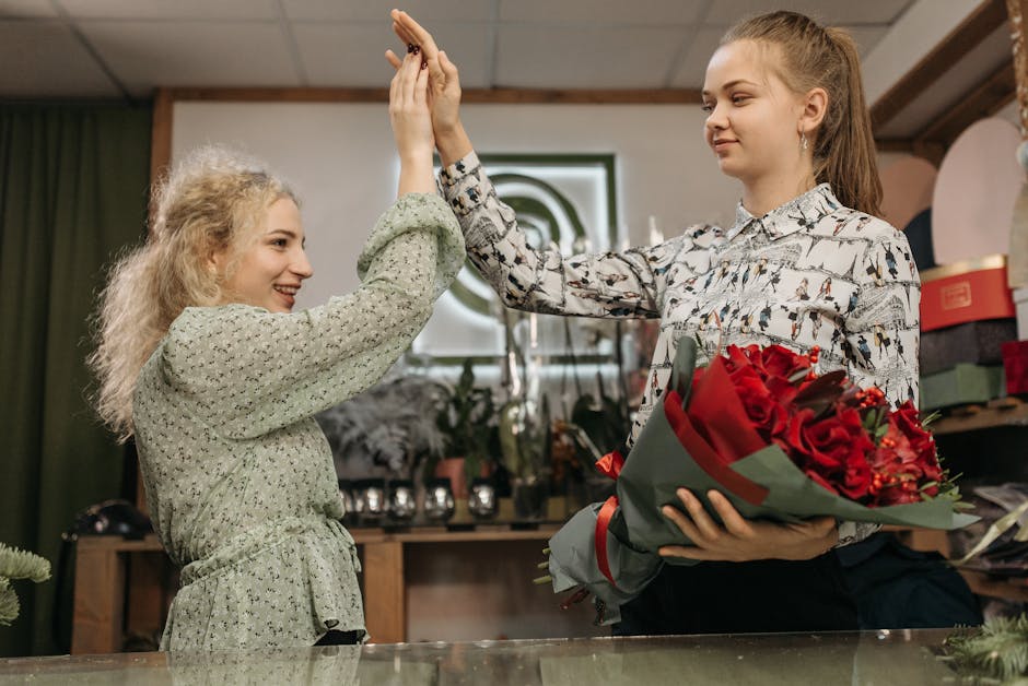 A woman with curly blonde hair wearing a light gray blouse with puffed sleeves is handing a tall bouquet of bright yellow sunflowers to a florist in a dark blazer inside a flower shop. The bouquet fea