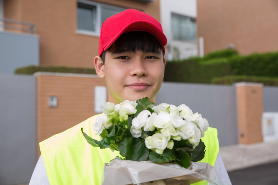 A person wearing a floral sleeve holds a bouquet of fresh pink and green flowers wrapped in light pink paper, extending towards a dark blue door with a decorative black wrought iron grille. The bouque
