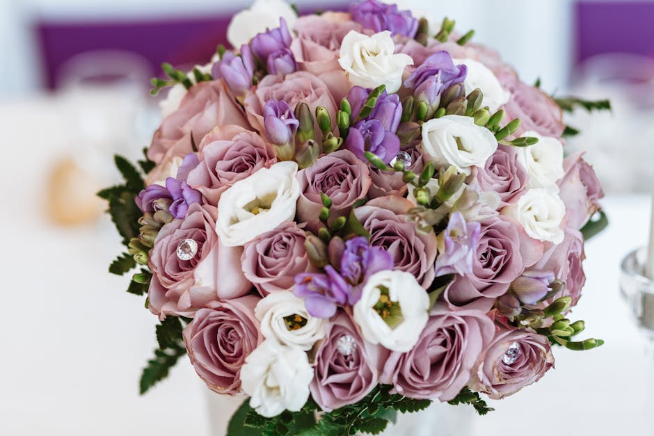 A close-up image of a floral arrangement featuring a bouquet of fresh roses in soft pinks and purples, accented with white eustoma flowers and small purple lisianthus. The bouquet is beautifully arran