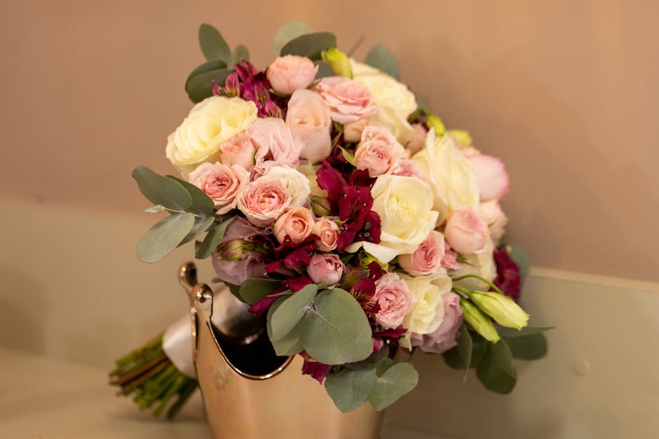 A close-up of a floral bouquet featuring a large, peach-colored rose at the center, surrounded by soft pink petals and accented by small clusters of blue and purple flowers. The arrangement is wrapped