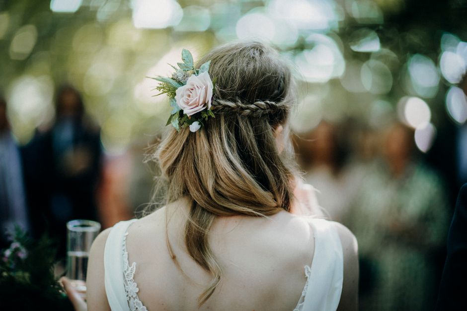 A woman with long, wavy hair styled in a loose braid, adorned with a floral hairpiece featuring soft pink roses, green leaves, and sprigs of lavender, is facing away in an outdoor setting. She is wear