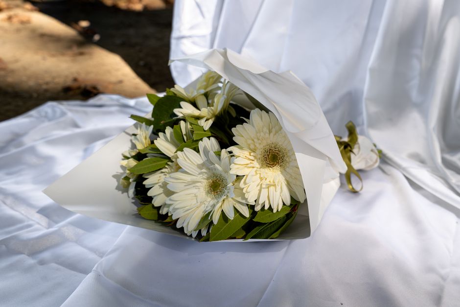 A bouquet of fresh white Gerbera daisies and white chrysanthemums arranged with green foliage, wrapped in white floral paper and tied with a gold ribbon. The bouquet is placed on a silky white fabric 