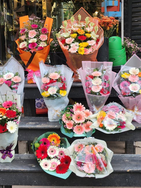 A display of various fresh flower bouquets arranged on a dark wooden outdoor table. The bouquets feature a mix of pink, white, yellow, and red daisies, gerbera daisies, and chrysanthemums, wrapped in
