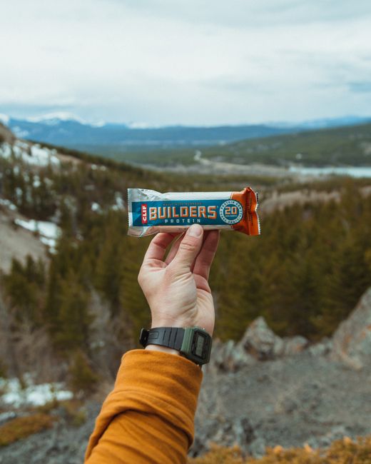 A person wearing a mustard-colored long-sleeve shirt and a black wristwatch is holding a protein bar labeled 'BUILDER'S PROTEIN' with '20g' in front of an outdoor landscape featuring rolling hills, sp