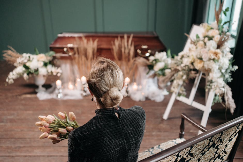 A woman arranging a floral tribute with a mixture of pale pink roses, white anemones, and greenery on a polished wooden casket during a funeral service. She is dressed in black, focusing attentively o