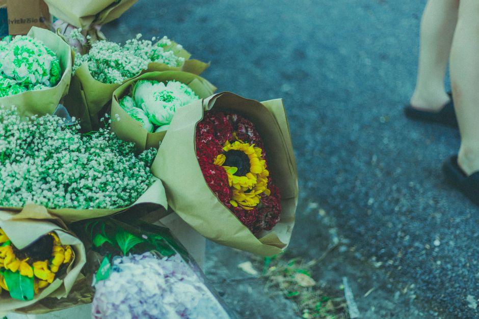 A collection of fresh floral bouquets arranged on a textured outdoor surface, featuring a variety of flowers including vibrant red carnations, yellow and purple daisies, and clusters of small white ba